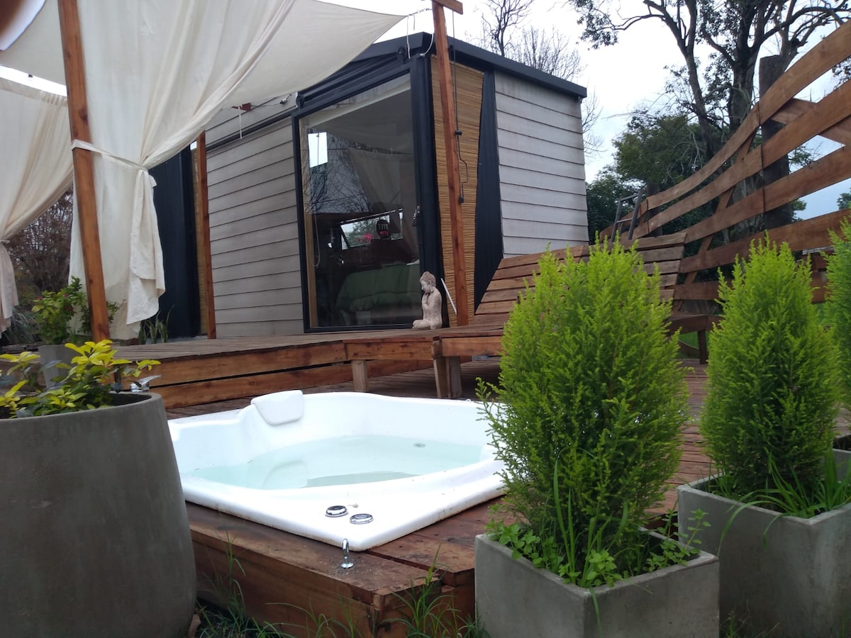 An inviting outdoor space features a private hot tub on a wooden deck, framed by planters with lush greenery. The structure in the background showcases large glass windows, allowing for natural light and views of the surrounding area.