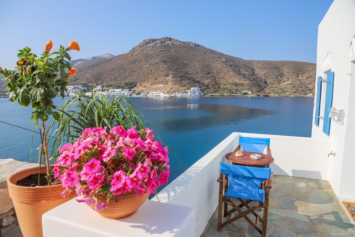 A scenic balcony overlooks the serene sea and surrounding hills. Bright pink flowers in a terracotta pot add a pop of color next to a small wooden table set with two blue chairs. The tranquil water reflects the clear blue sky.
