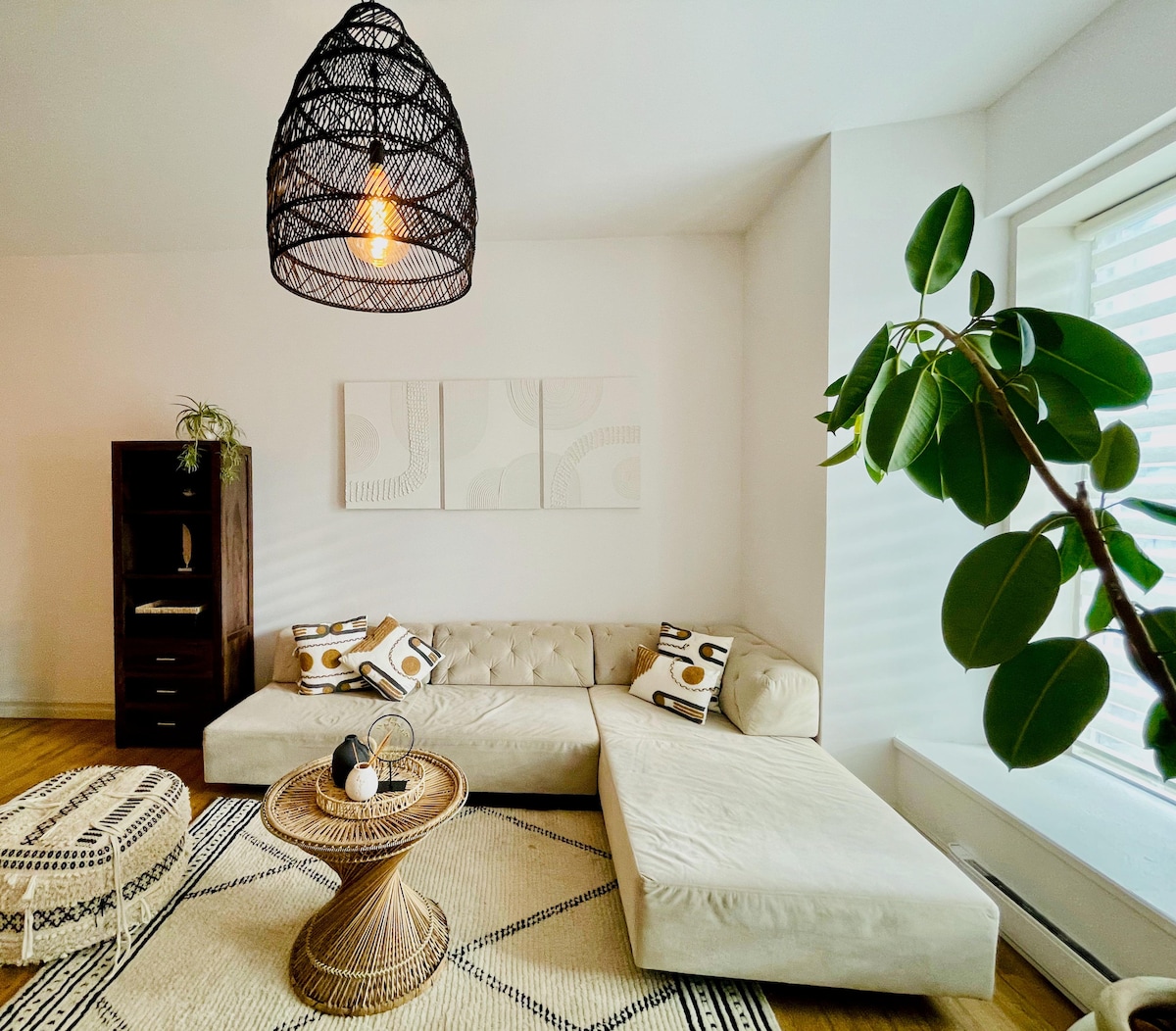 A light and airy living space is presented with a sectional sofa upholstered in cream fabric. A round coffee table, crafted from natural materials, is centered on a patterned area rug. Soft light filters through the large window adorned with sheer blinds, highlighting the minimalist decor.