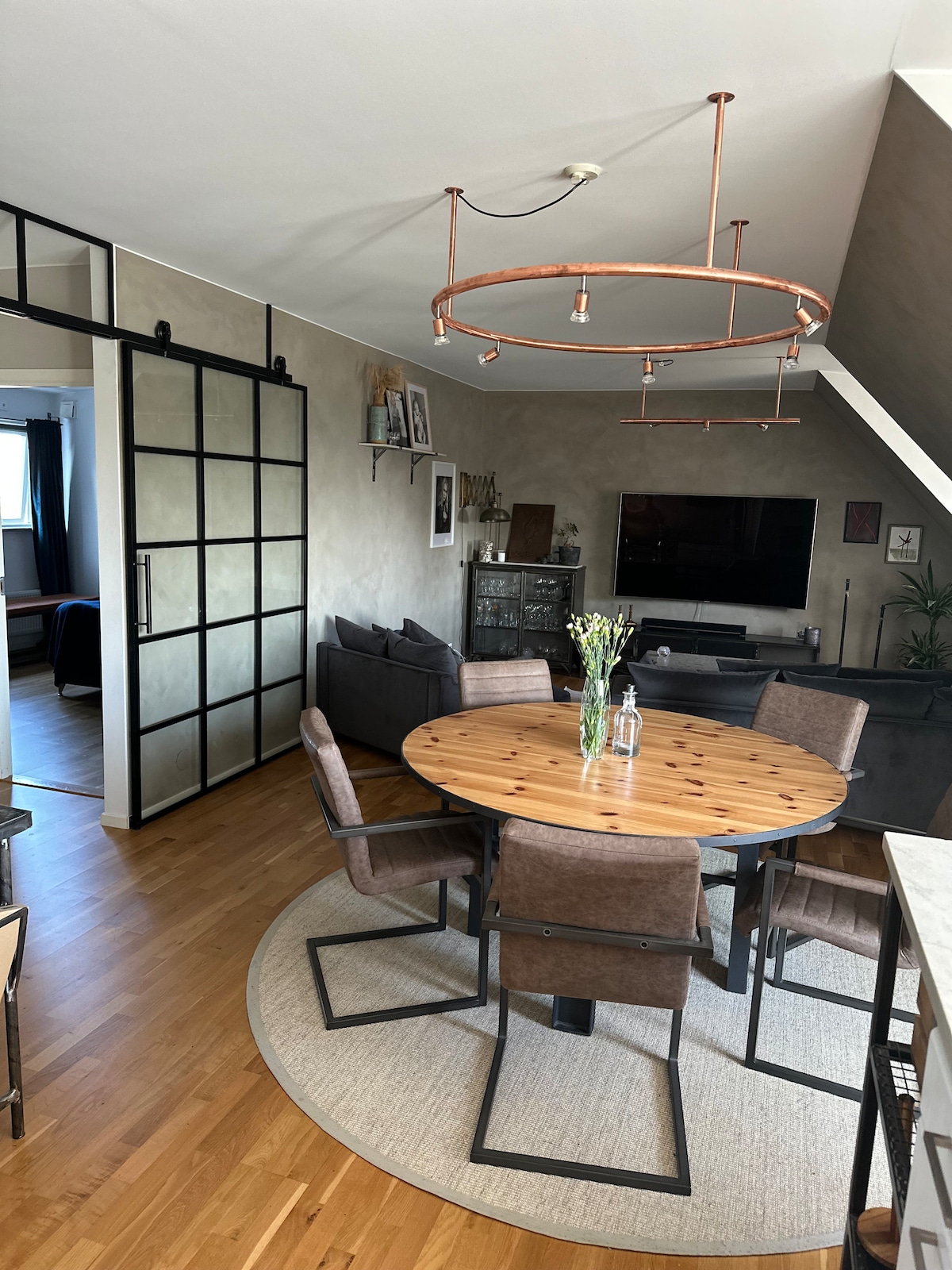 A dining area is shown with a round wooden table surrounded by four metal-framed chairs. A light fixture hangs above the table, and a rug is placed underneath. A black sofa and a large television can be seen in the background, along with decorative wall art.