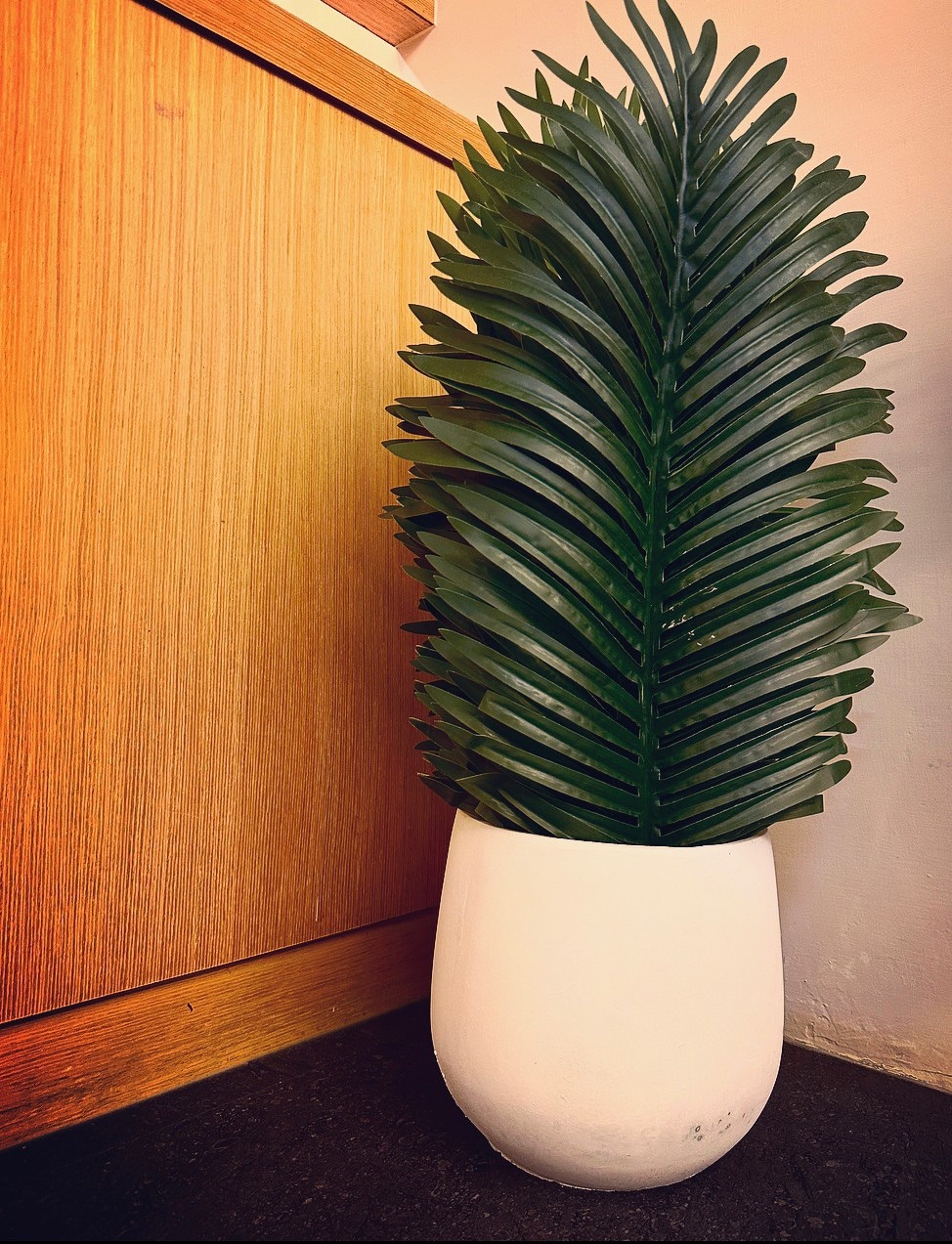 A tall, leafy faux plant is displayed in a white planter. The planter rests against a wooden wall, providing a natural touch to the indoor space. The plant's vibrant green leaves create a contrast with the earthy tones of the surrounding decor.