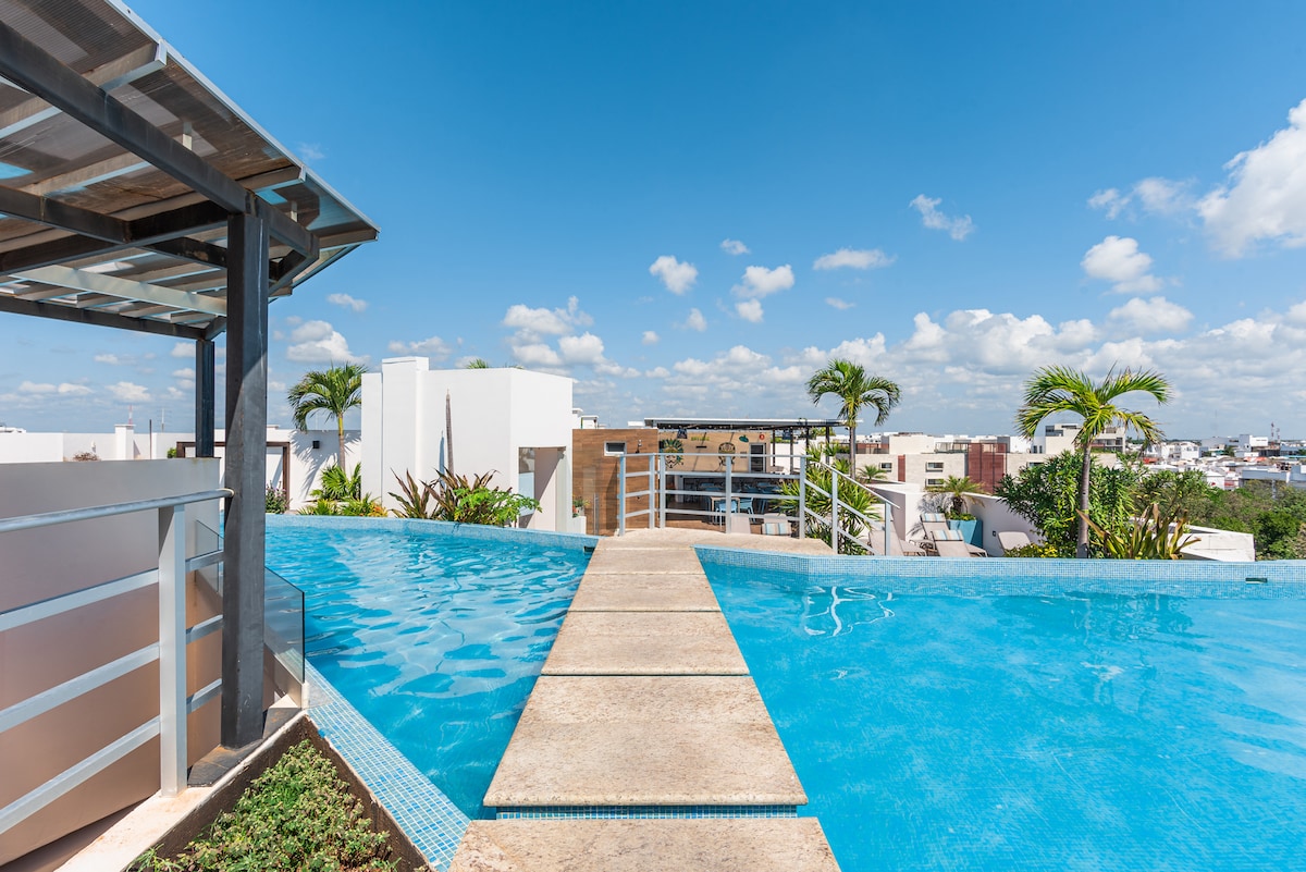 An inviting rooftop pool is showcased with a smooth stone pathway leading across its surface. Lush greenery and palm trees are visible in the surroundings, with a bright sky dotted with soft clouds, creating a refreshing atmosphere.