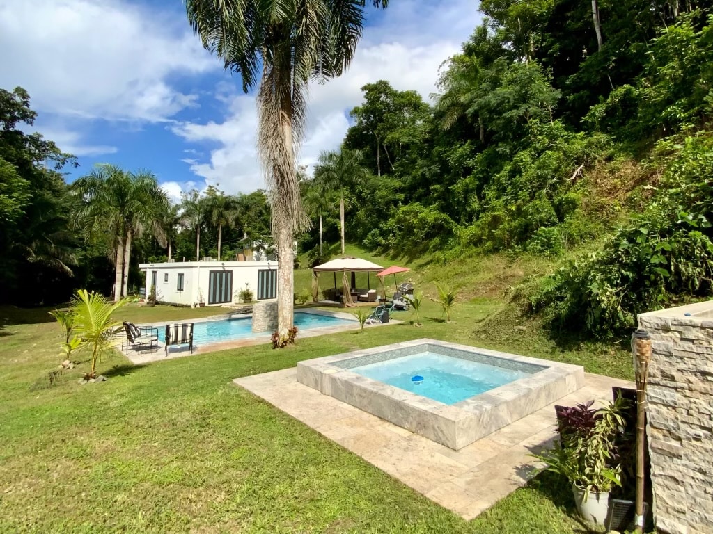 A spacious outdoor area is shown, featuring a large pool and a hydromassage jacuzzi, both surrounded by lush tropical vegetation. A shaded seating area with an umbrella is present nearby, contributing to the serene and natural setting.