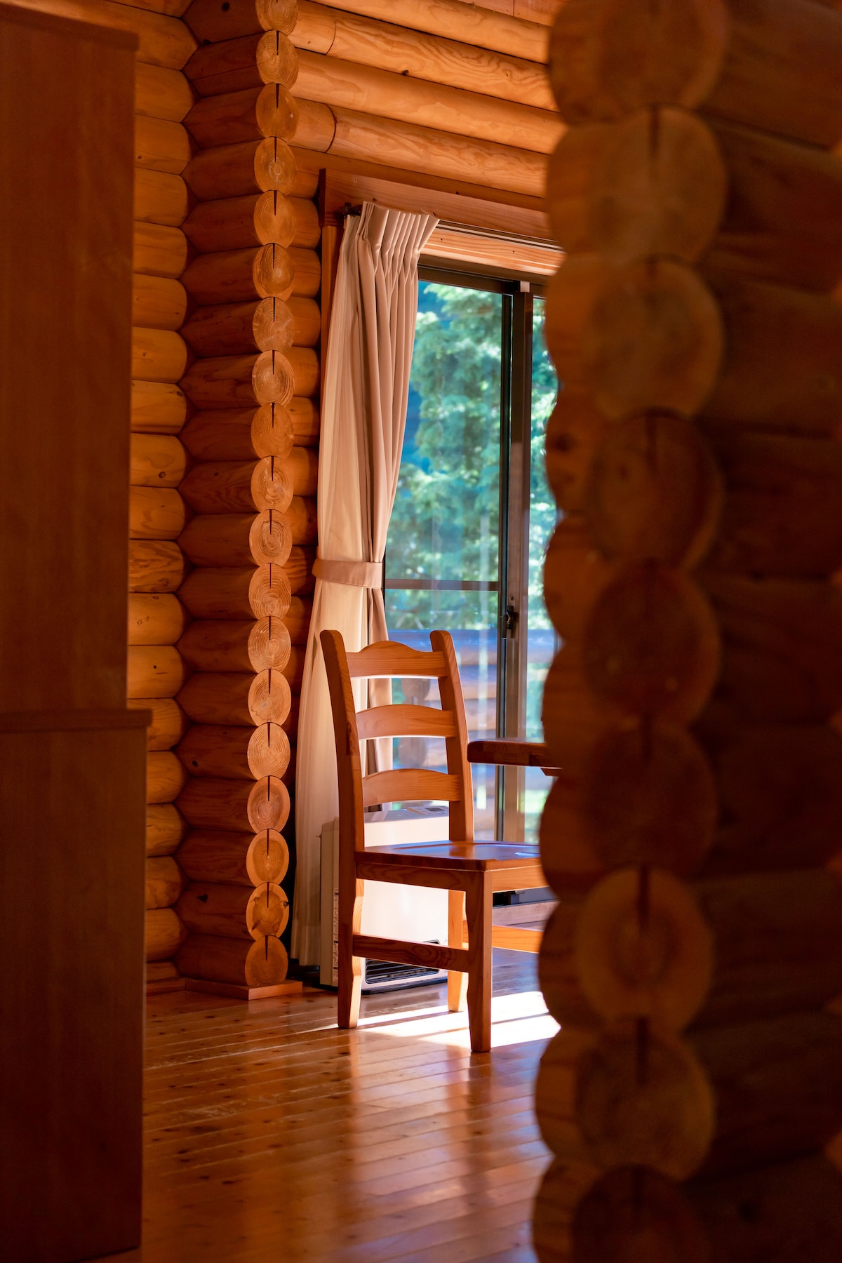 A wooden chair is positioned beside a window, inviting natural light into the space. The rustic log walls add warmth to the interior, while a glimpse of greenery is visible through the glass door, enhancing the connection with the surrounding nature.