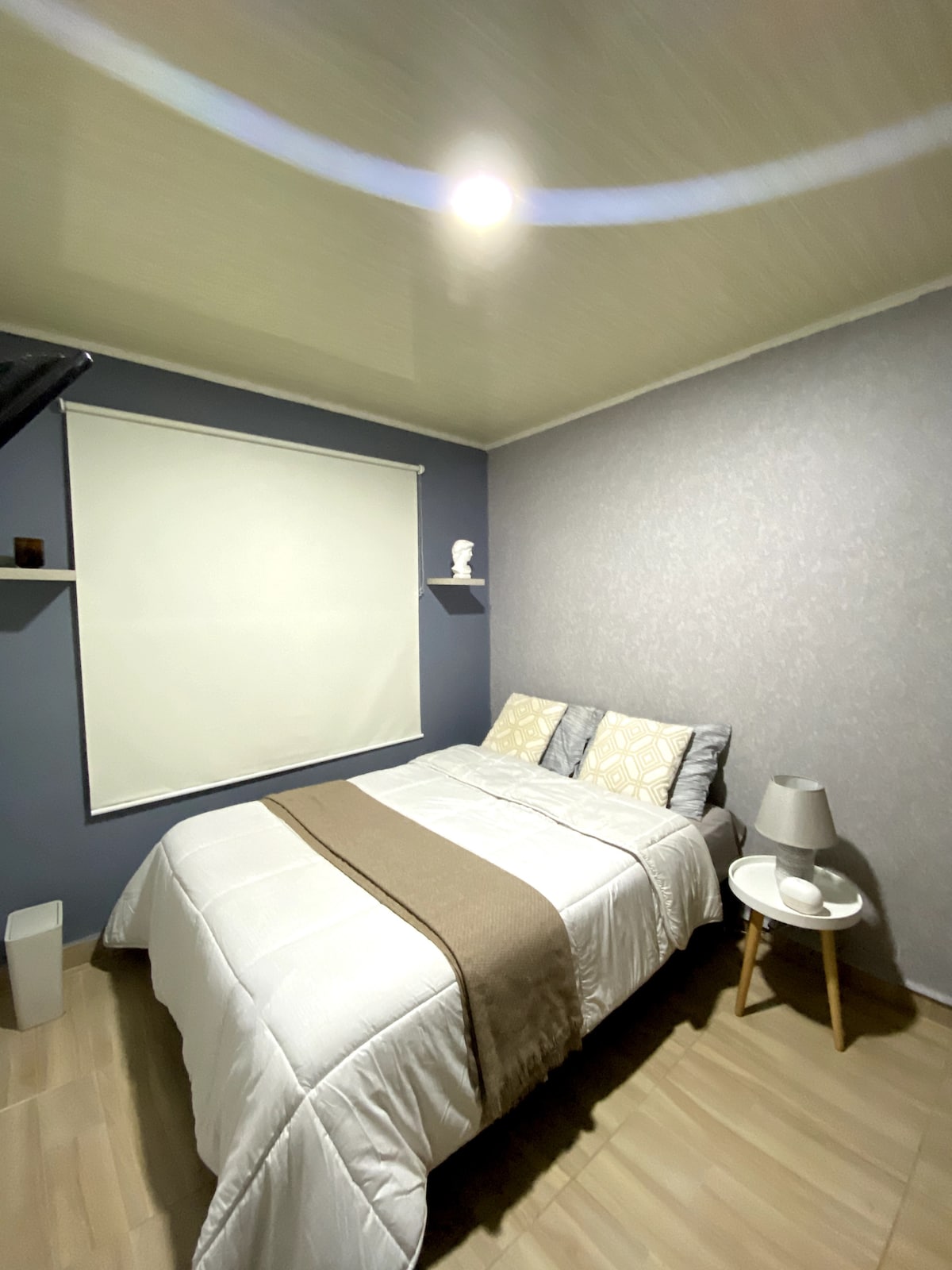 A serene bedroom features a spacious bed covered with a light blanket and accented by decorative pillows. A small round table with a lamp is situated beside the bed. A subtle wall-mounted shelf holds a decorative item, and a roller blind covers the window.