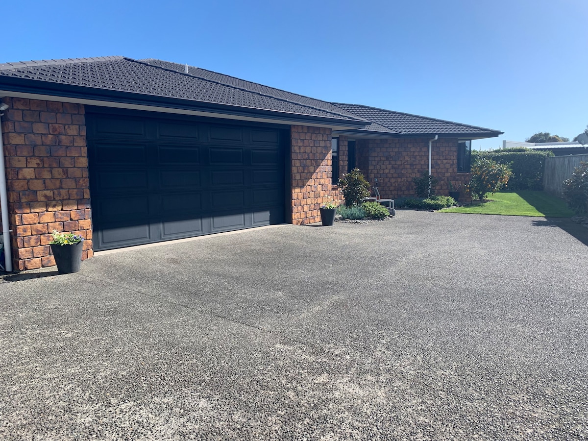 A modern home with a textured brick exterior is set against a clear blue sky. The driveway is spacious, leading to a large two-car garage. Neat landscaping with potted plants frames the entrance, enhancing the inviting appeal of the property.
