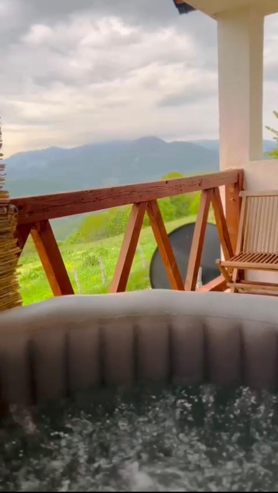 A hot tub with bubbling water is positioned on a balcony. Wooden railings frame the space, providing a view of the surrounding mountains and lush greenery. A pair of wooden chairs are placed nearby, inviting relaxation amidst a tranquil outdoor setting.