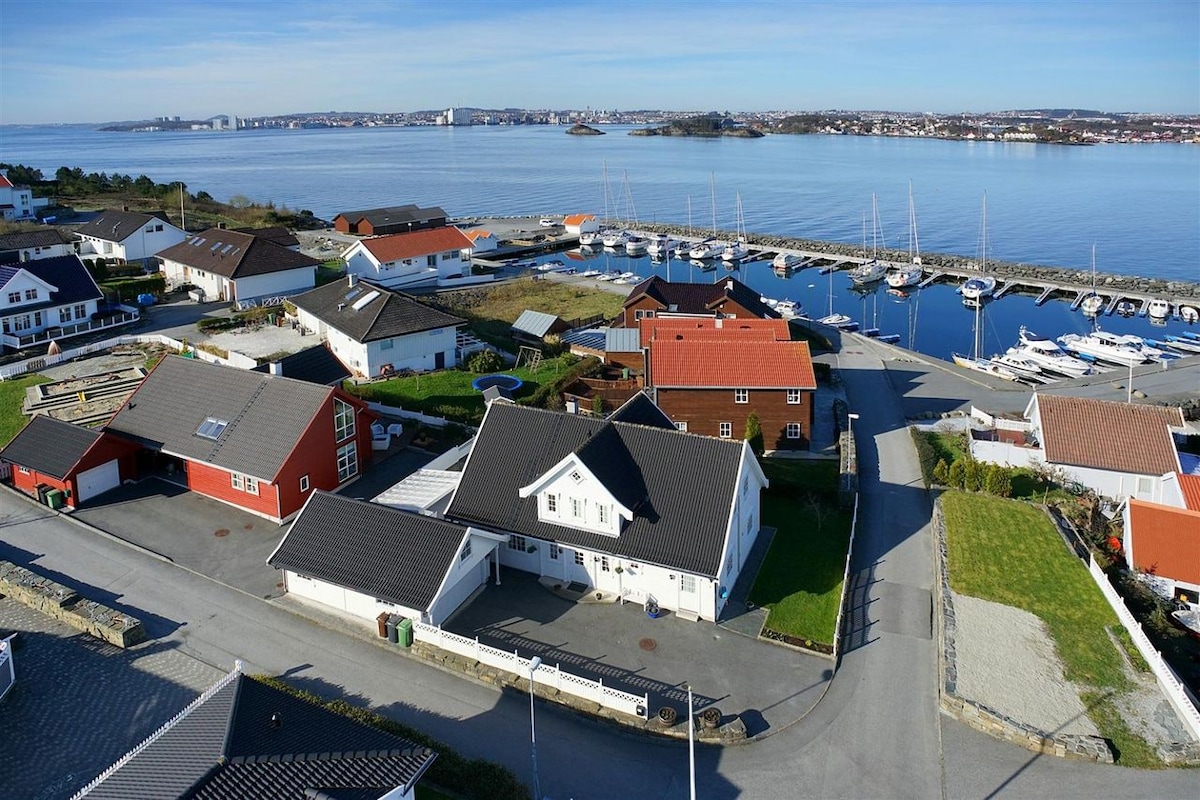 Aerial view of a serene coastal neighborhood featuring a marina with several boats, surrounded by residential homes. The well-maintained roads and green spaces reflect a peaceful atmosphere, while the distant views of the water and distant buildings enhance the idyllic setting.