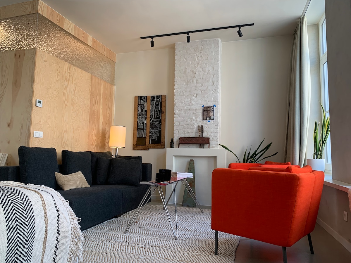 A comfortable living area features a dark sectional sofa accompanied by a bright orange armchair. A crystal-like wall adds texture, while a soft rug anchors the space. Natural light enters through large windows, and decorative plants enhance the vibrant atmosphere.