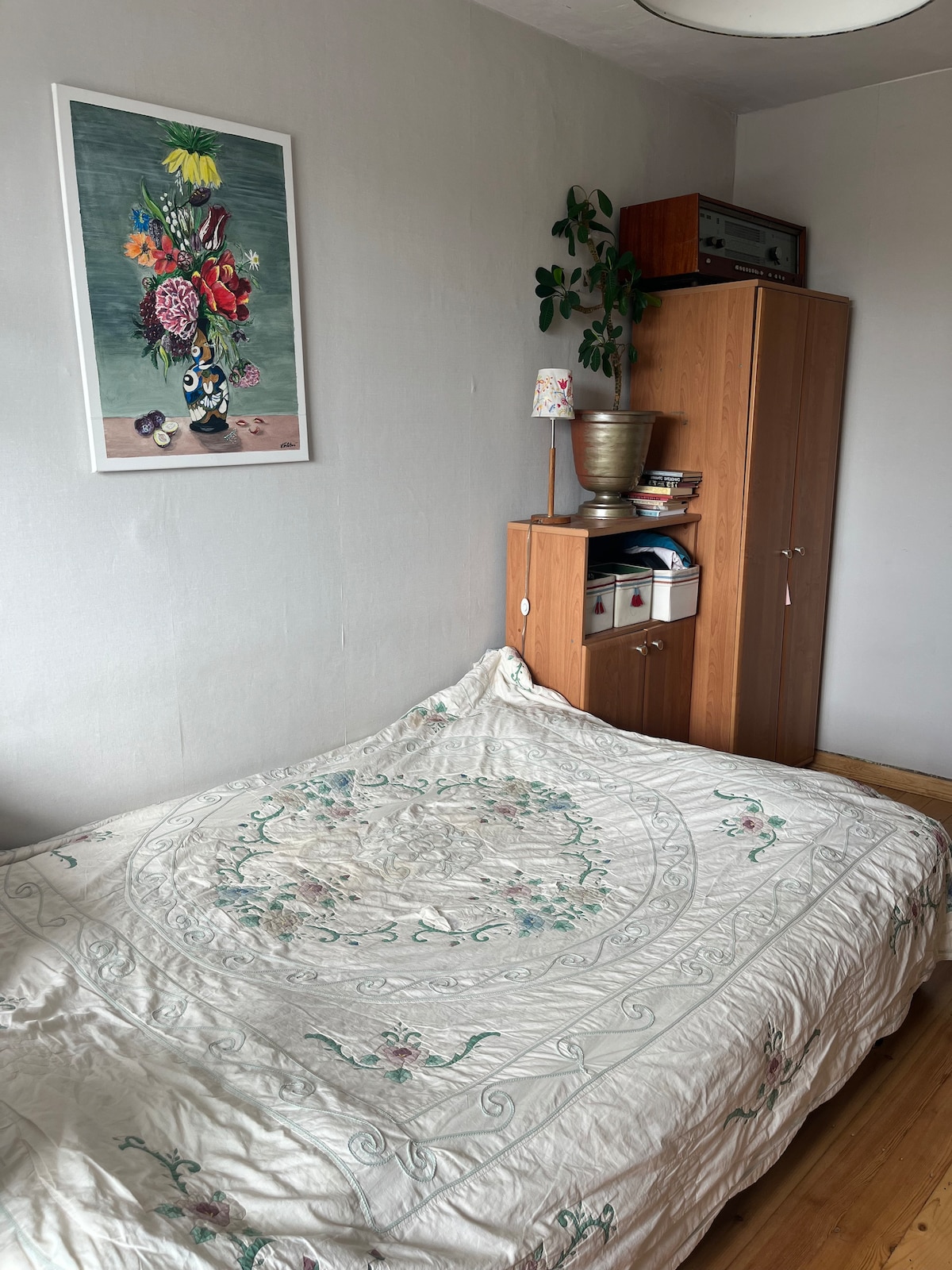 A spacious bed is covered with a light-colored quilt featuring floral embroidery, positioned against a soft grey wall. A decorative plant and a lamp sit atop a wooden nightstand, while a cabinet with storage bins stands nearby, contributing to the room's functionality.