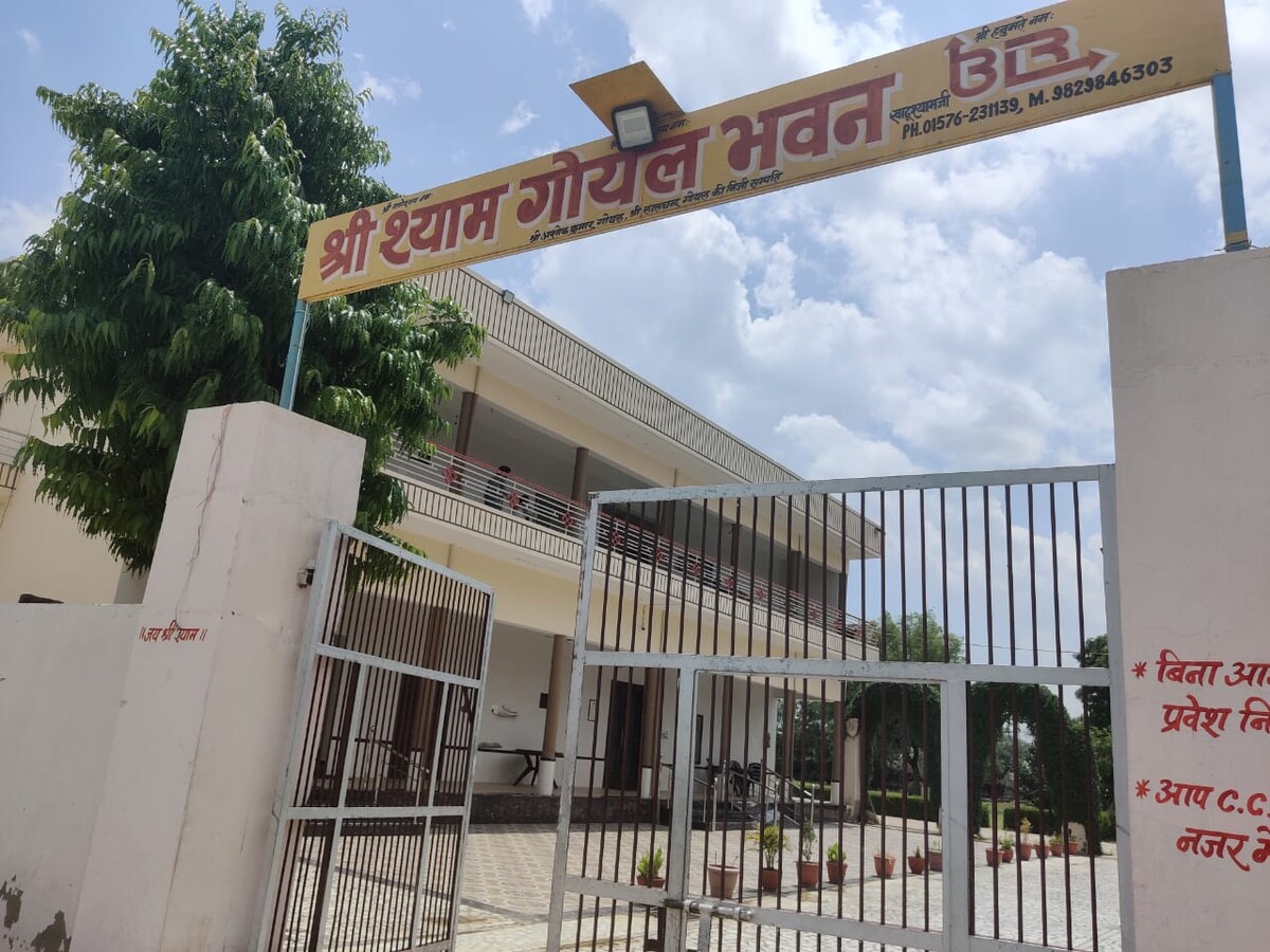 A welcoming entrance is presented, featuring a gated entry and a well-maintained exterior. The building shows a two-story structure with a clean facade and balcony details. Surrounding greenery adds a natural touch, while the sky overhead is partly cloudy.