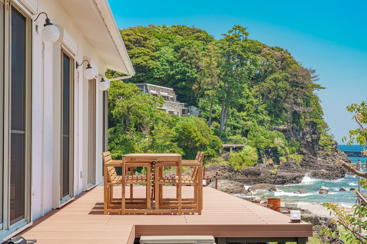 An outdoor deck features a wooden dining table surrounded by chairs, overlooking a scenic view of the coastline and lush greenery. The ocean can be glimpsed in the distance, providing a connection to the natural surroundings.