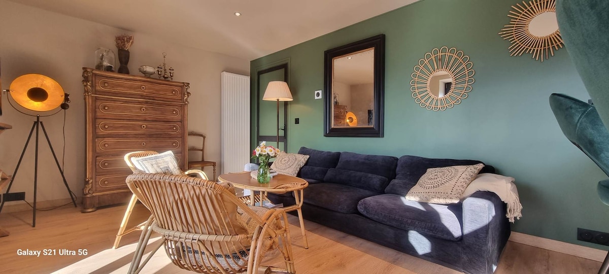 A cozy living area features a dark blue sofa and a light wooden coffee table, surrounded by a rattan chair. Green walls are complemented by a decorative mirror and wall art, while warm lighting creates a relaxed ambiance.