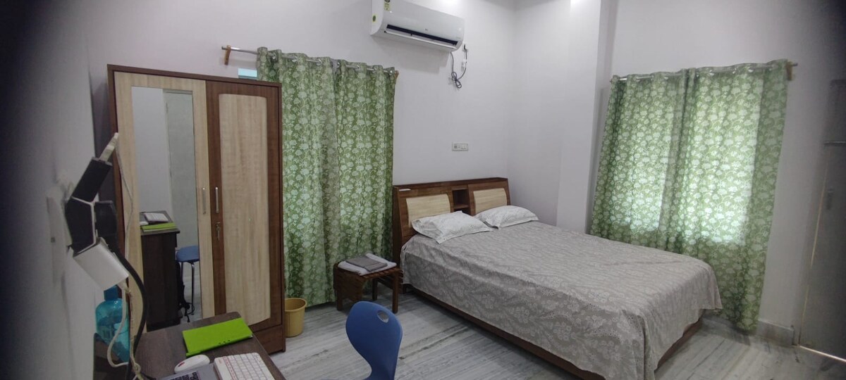 A spacious bedroom is furnished with a double bed featuring a white and grey patterned bedspread. Light green curtains frame the window, allowing natural light to enter. A small desk is positioned nearby, with a computer, and a simple wardrobe is present against the wall.
