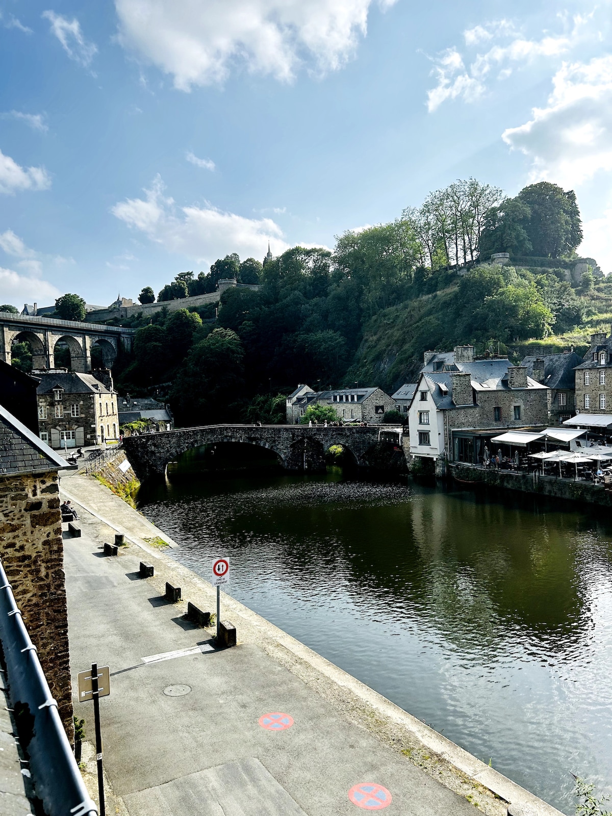A serene view of a river, framed by charming stone buildings that line its banks. A historic stone bridge crosses the water, while a lush hillside rises in the background, dotted with trees and greenery. Soft clouds drift across a blue sky.