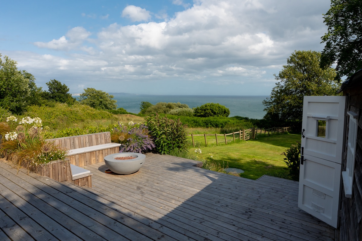 A spacious wooden deck extends outdoors, featuring a circular fire pit at its center. Comfortable seating is arranged nearby, surrounded by lush green grass and blooming flowers. The ocean can be seen in the background, with trees framed along the horizon under a partly cloudy sky.