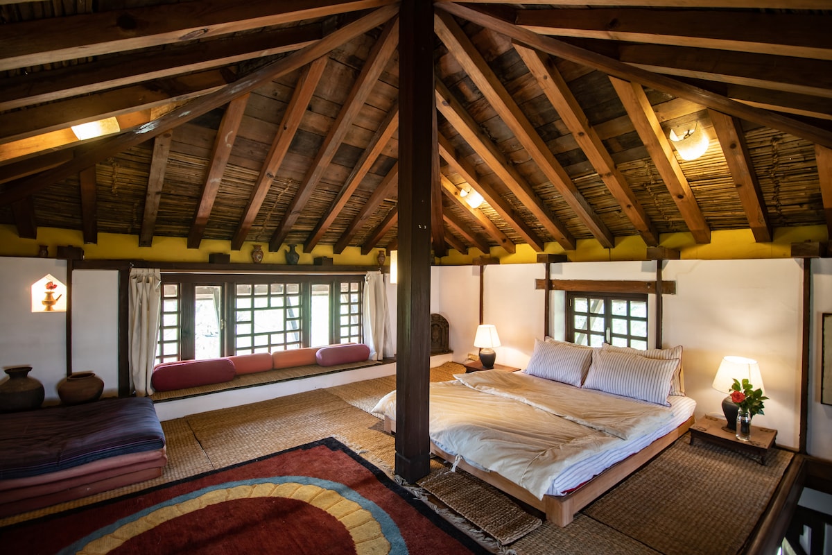 The spacious interior features traditional wooden beams and a high, sloping ceiling. A large bed is positioned alongside a low table, with natural light filtering through the windows. Soft mats and cushions are arranged for sitting, enhancing the cozy ambiance of the room.