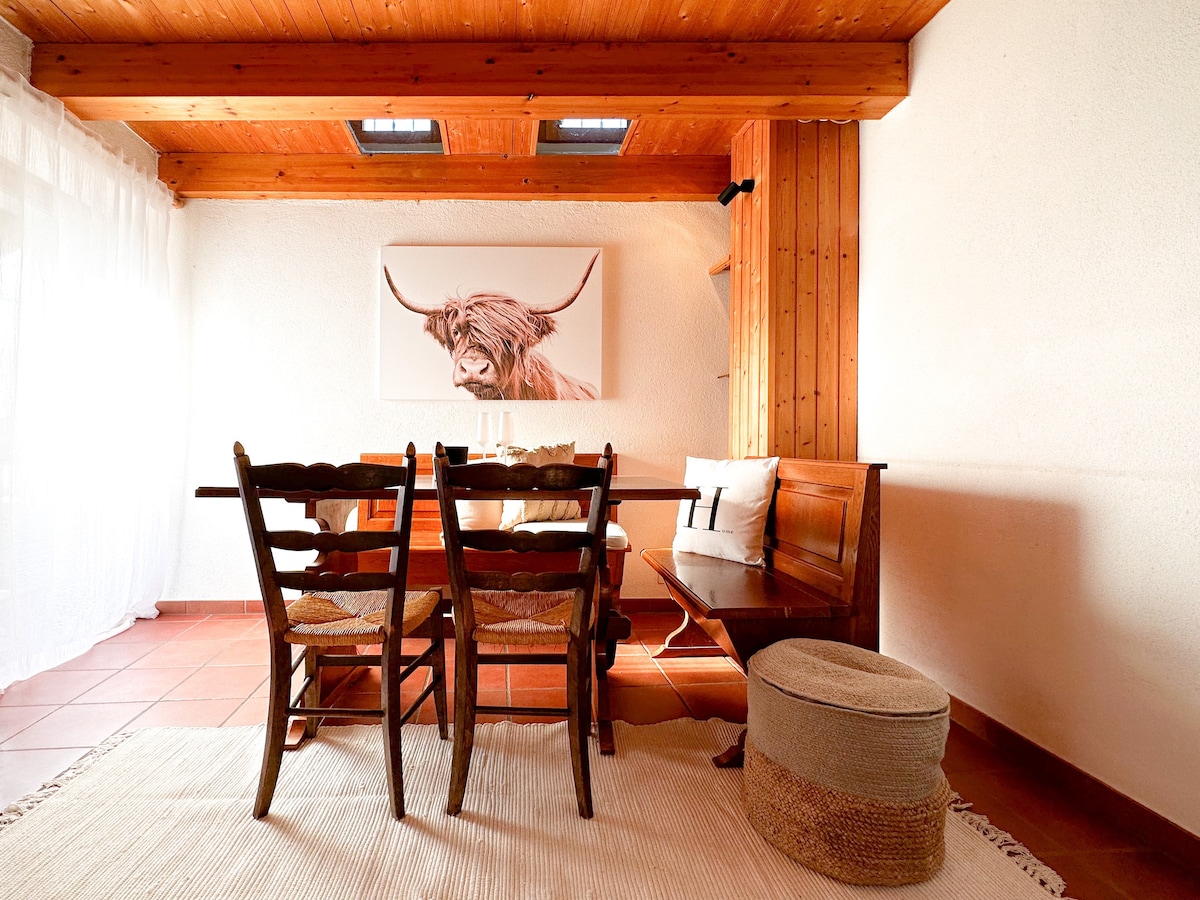 A wooden dining area features a table surrounded by four chairs. A soft-textured rug lies underneath, while a large artwork depicting a Highland cattle is mounted on the wall. Natural light filters through sheer curtains, enhancing the cozy ambiance.