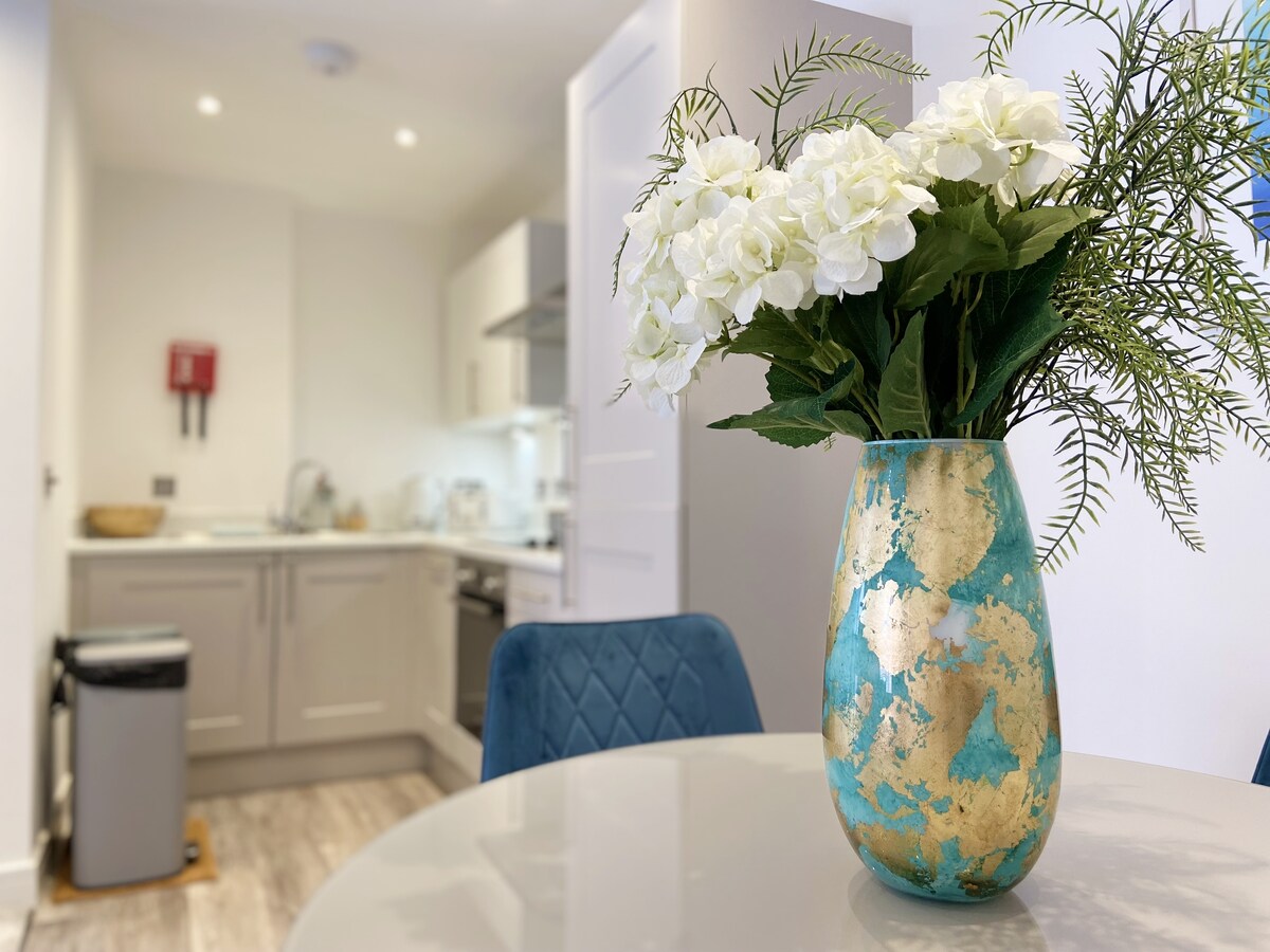A dining area is presented with a round table, adorned by a decorative vase filled with white flowers. The kitchen is visible in the background, featuring modern appliances and cabinetry. Soft lighting enhances the welcoming ambiance of the open-plan space.