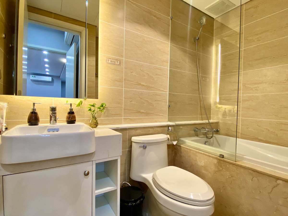 The modern bathroom features a clean design with a bathtub and separate shower. A sleek sink is complemented by minimalist cabinetry for storage. Natural light is provided through a glass partition, enhancing the spacious feel of the area.