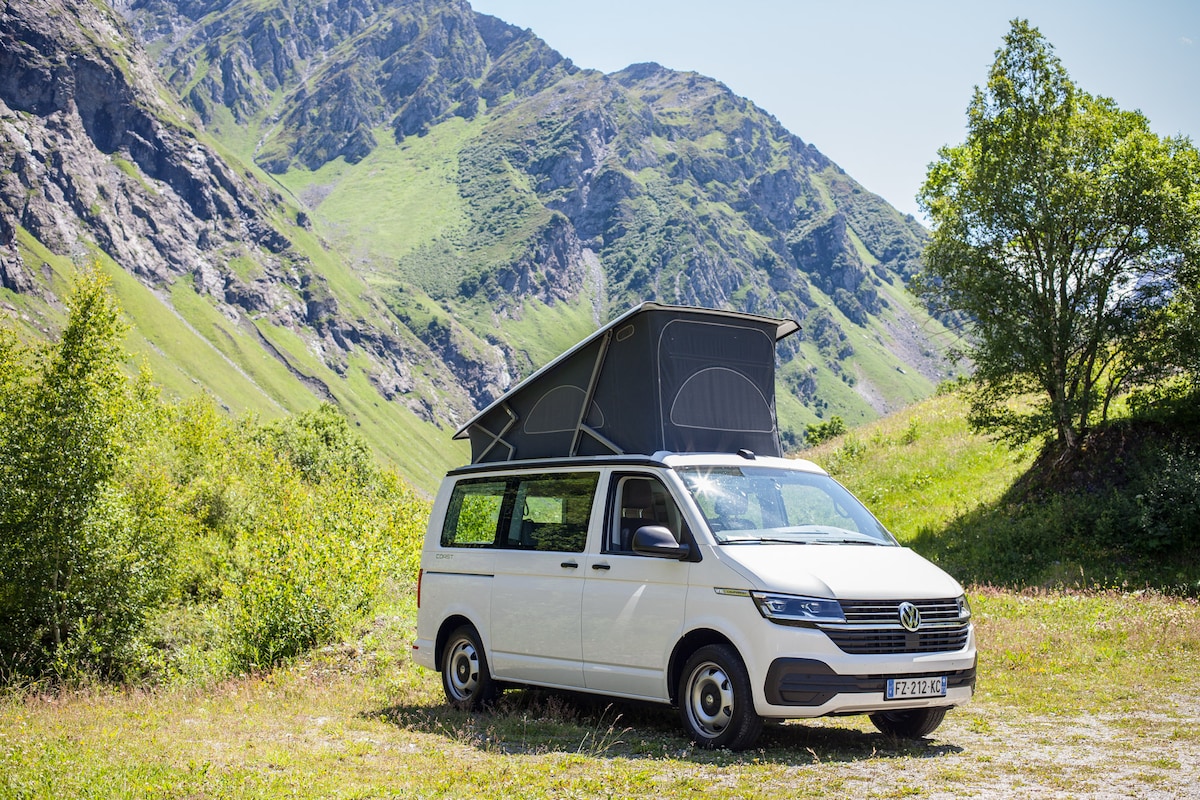 A well-equipped van is parked in a lush green meadow, surrounded by towering mountains. The elevated roof adds extra space, while the bright sunlight reflects off the vehicle's exterior, inviting adventure in a serene natural setting.