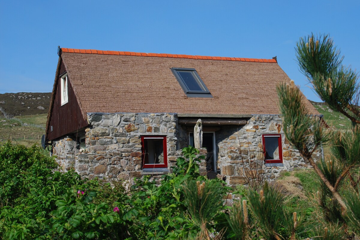 A rustic cottage is depicted, featuring a stone exterior with a thatched and sloped roof. Bright red window frames contrast against the natural stone. Lush greenery surrounds the building, while a clear blue sky is visible above, enhancing the peaceful ambiance of the rural setting.
