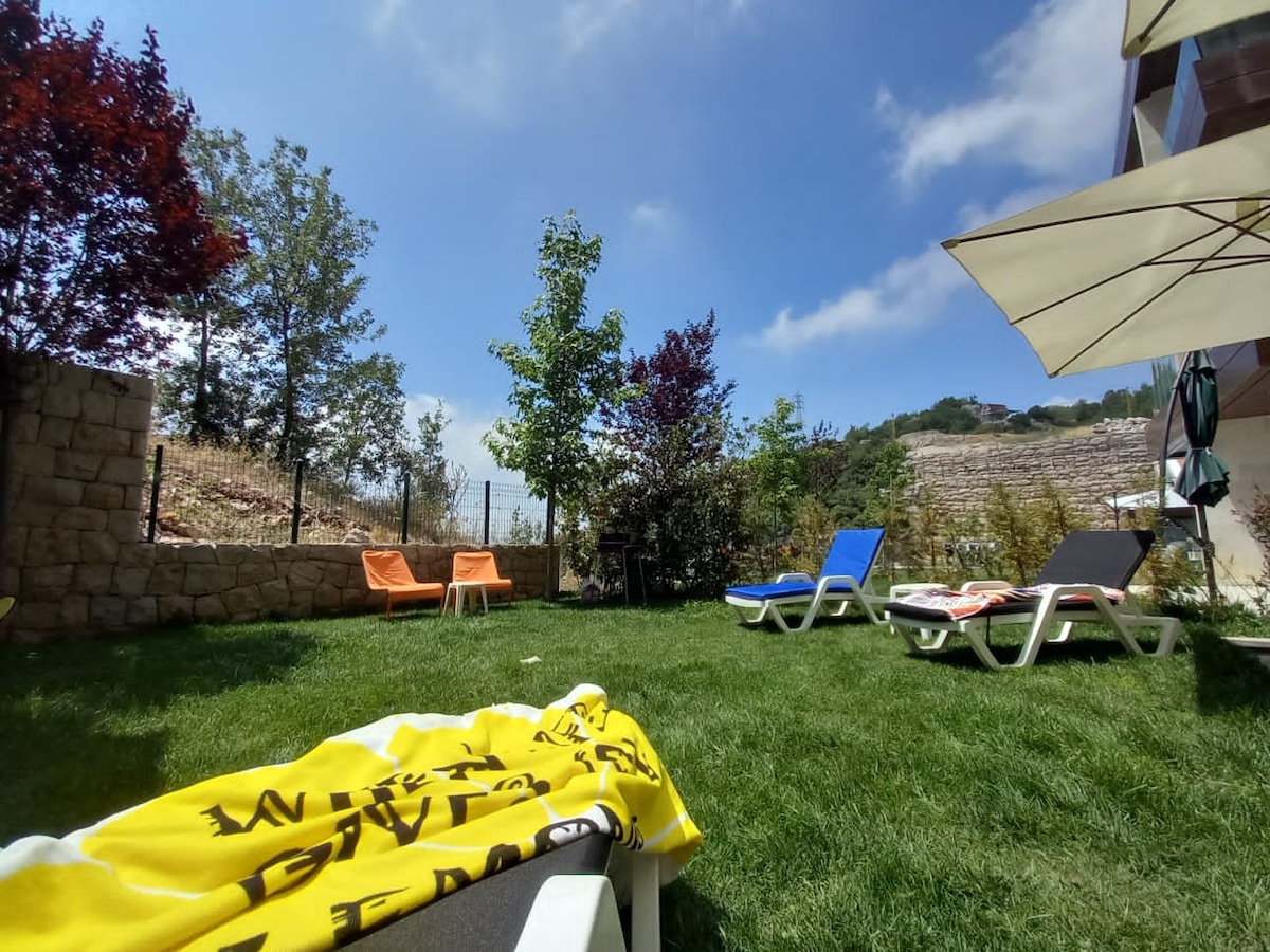 A grassy garden area is surrounded by trees and shrubs, with colorful lounge chairs positioned on the lawn. A sunny blue sky stretches overhead, and a stone wall marks the boundary of the garden, offering a peaceful outdoor space.