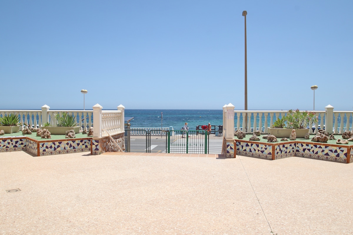 A spacious outdoor area offers a clear view of the sea, with a decorative tiled border and potted plants lining the space. The calm waters reflect the sky, while a gated entrance provides access to the beach. Natural light illuminates the scene.