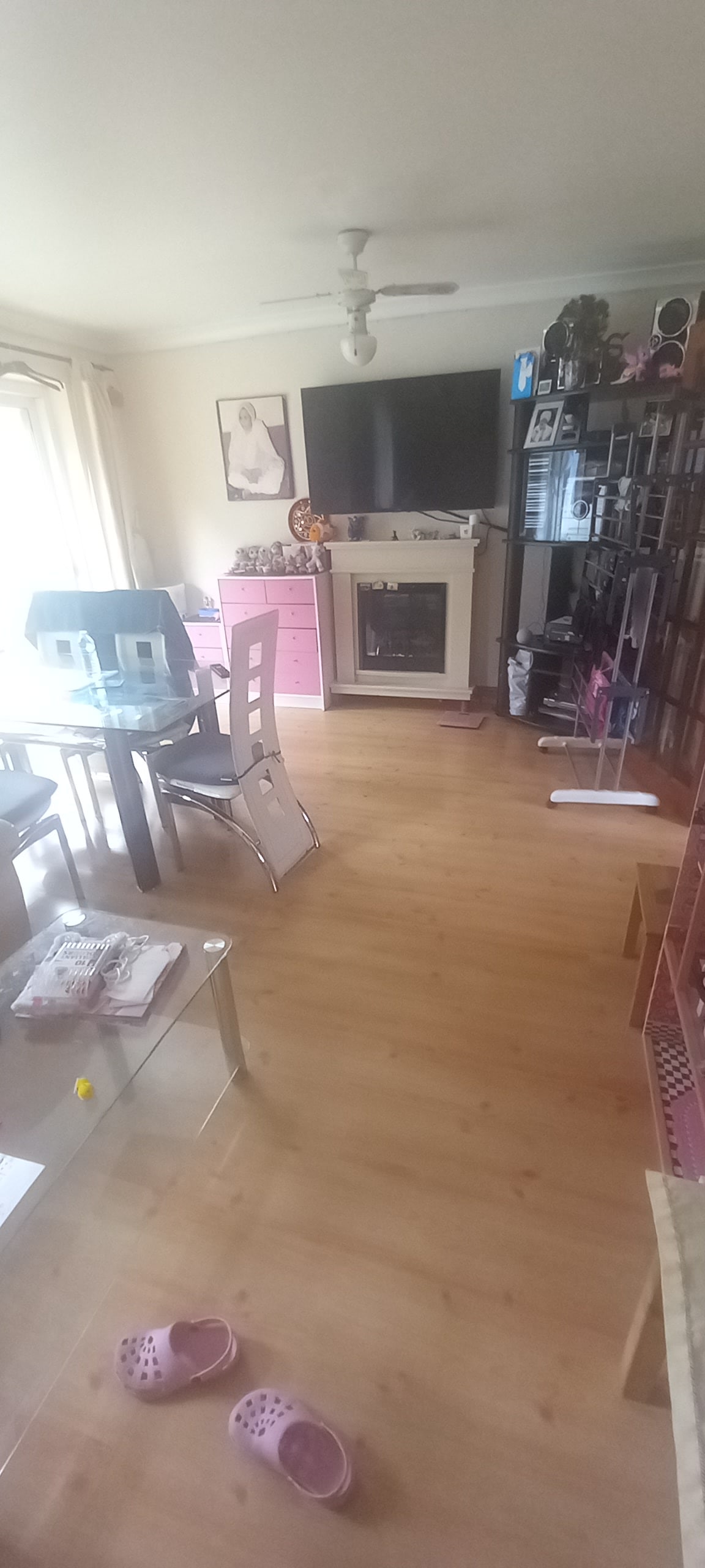 A living area features a glass dining table and metal chairs, with a fireplace and television unit on one side. Light-colored wooden flooring runs throughout the space, complemented by a variety of decorative items and a gentle natural light filtering through the curtains.