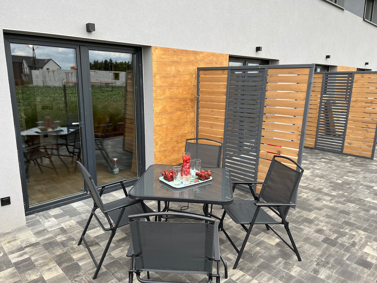 An outdoor seating area features a rectangular table surrounded by six black chairs. The space is bordered by wooden screens for privacy, with large glass doors reflecting the surrounding greenery.