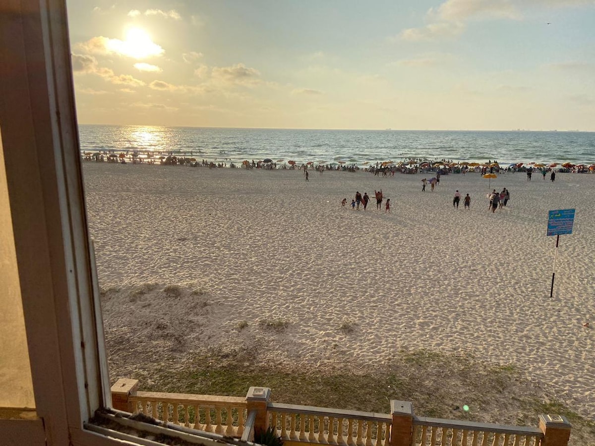 A tranquil view of a sandy beach at sunset is captured from a window, highlighting the expansive sea and people enjoying the shoreline. The warm sunlight reflects off the water's surface, creating a serene atmosphere, while beach umbrellas can be seen in the distance.
