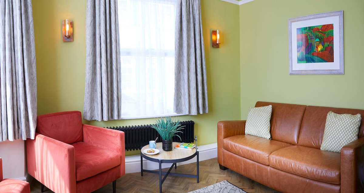 A cozy living area is highlighted by a tan leather sofa paired with two red accent chairs. Soft lighting from wall sconces complements the bright green walls. A circular coffee table holds a small plant and snacks, while artwork adds a touch of color to the space.