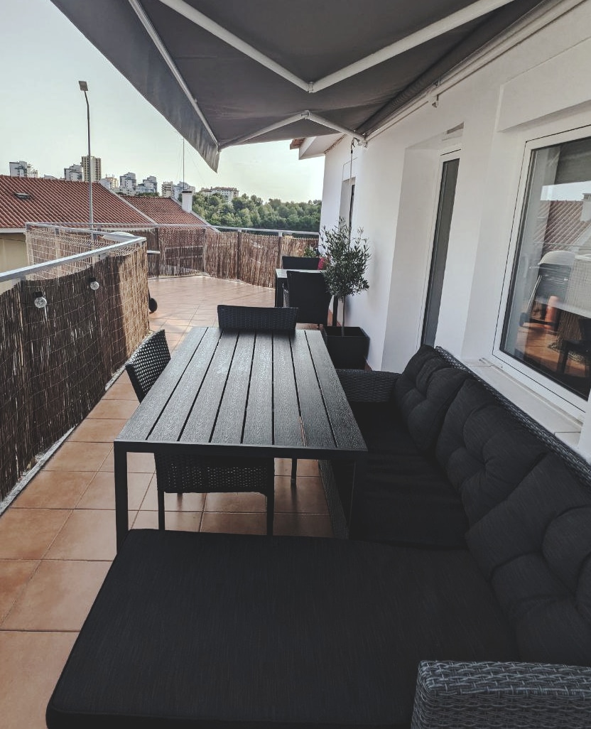 A spacious terrace is furnished with a black outdoor dining table and comfortable seating. Potted plants are placed strategically along the railing, and an awning provides shade, creating a relaxed outdoor space with views of the surrounding area.
