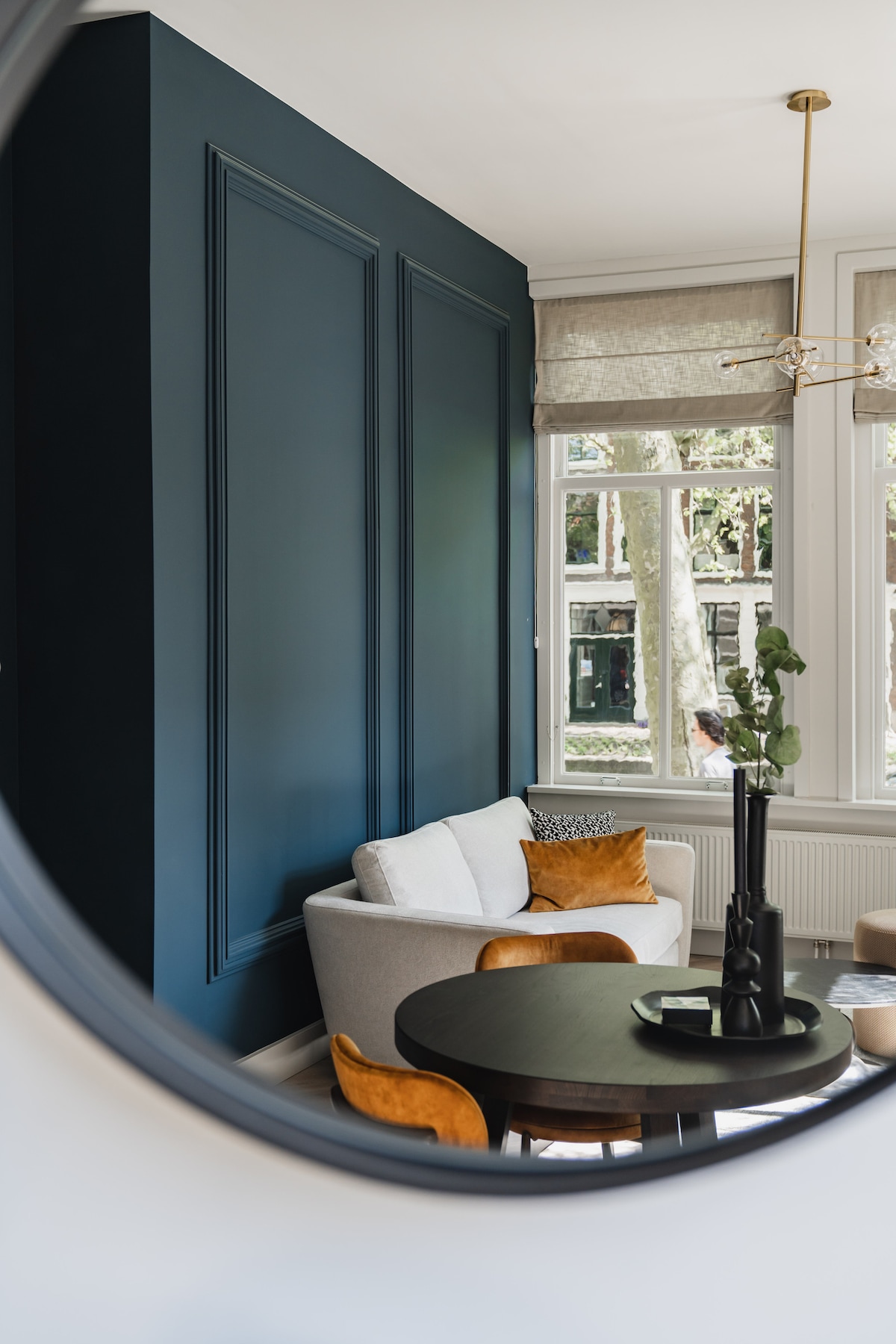 The living area is designed with a dark blue accent wall and features a light-colored sofa adorned with decorative cushions. A round dining table is situated nearby, accompanied by four chairs. Natural light enters through large windows, enhancing the inviting atmosphere.
