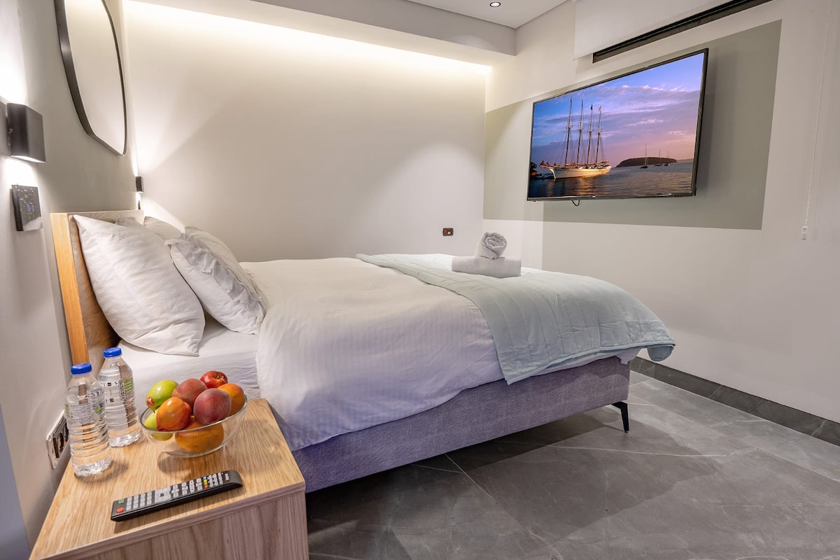 A well-lit bedroom features a spacious bed dressed in crisp linens, complemented by a light blue blanket. A flat-screen television is mounted on the wall, while a bedside table holds a bowl of fresh fruit and bottles of water. The room has a modern design with tiled flooring.