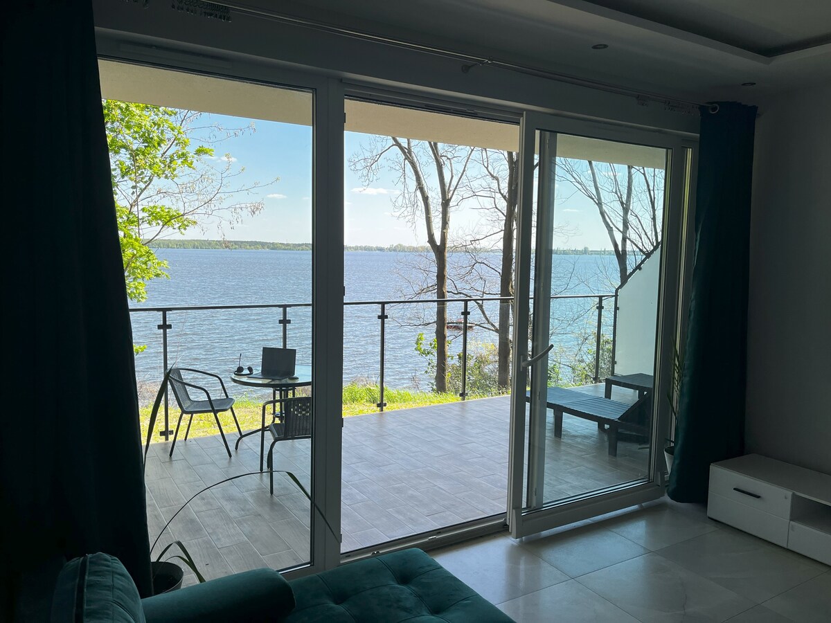 Large glass doors open to a view of a serene lake, framed by green trees. A spacious terrace with outdoor seating is visible, inviting relaxation. The interior features a cozy seating area with a blue sofa, providing a comfortable spot to enjoy the view.