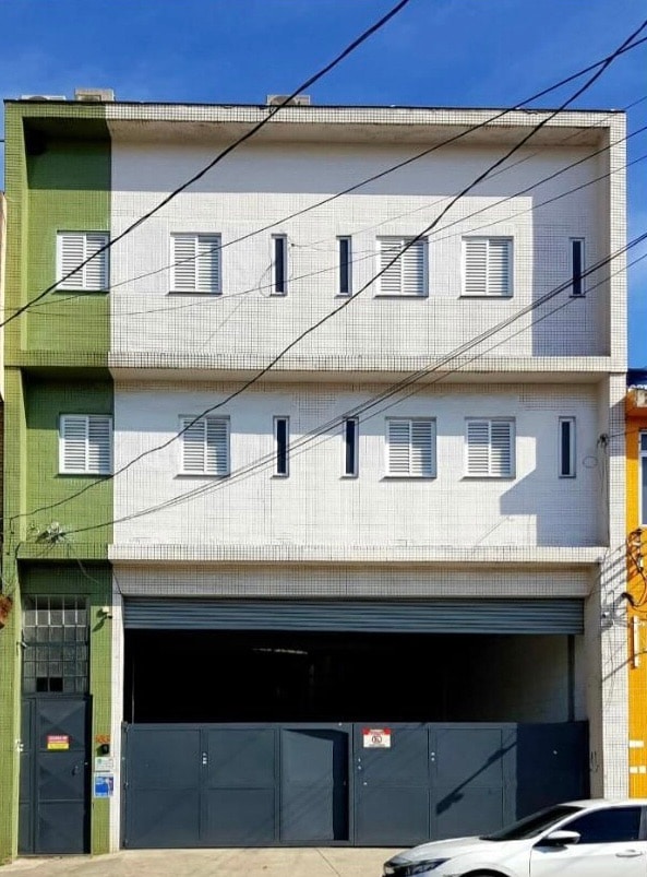 The exterior of a three-story building is presented, featuring a white facade with green accents. Multiple shuttered windows are evenly spaced across each level, and a large garage door is visible at the ground level. The sky above is clear and bright.