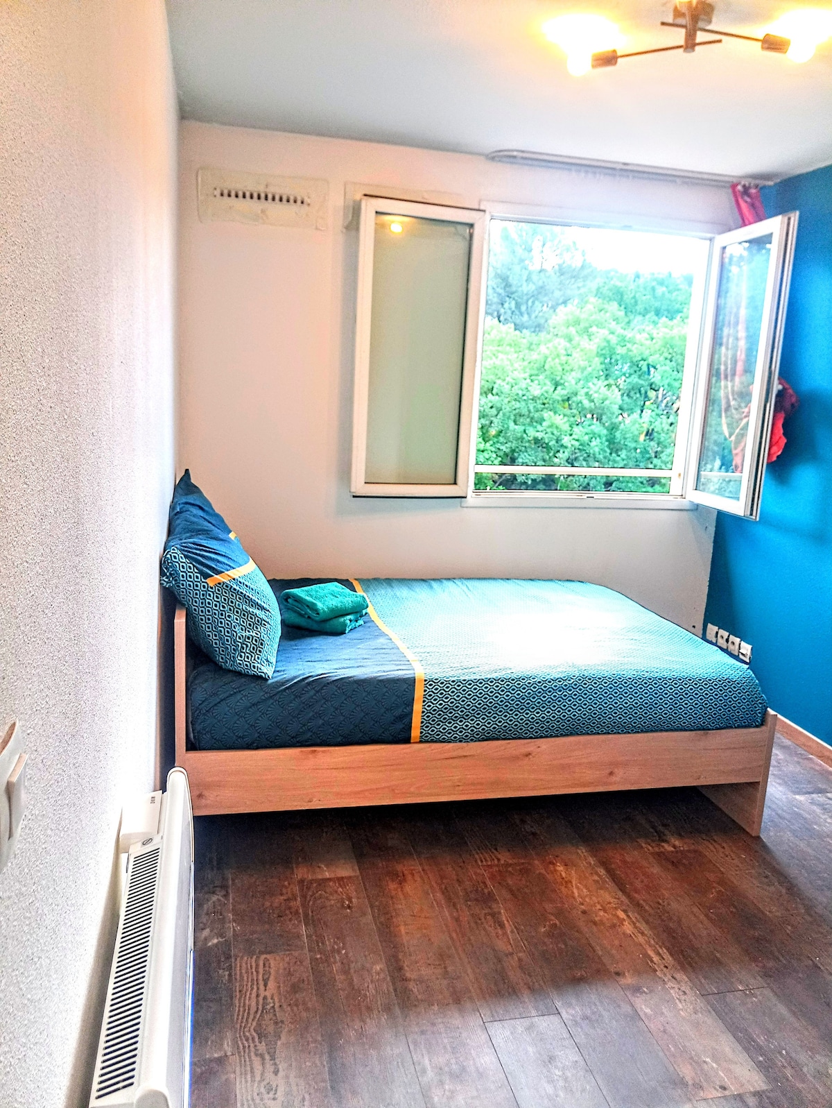 A cozy sleeping area features a wooden frame bed dressed in teal bedding, accompanied by a decorative pillow. Large windows brighten the room, offering views of the greenery outside. A radiator is visible along the wall, and a light fixture overhead provides illumination.