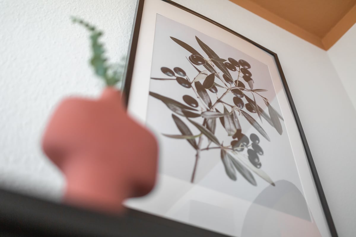 A framed artwork featuring a black and white image of olive leaves is displayed on a wall. A terracotta vase with a small green plant is positioned in the foreground, providing a touch of nature to the modern decor.