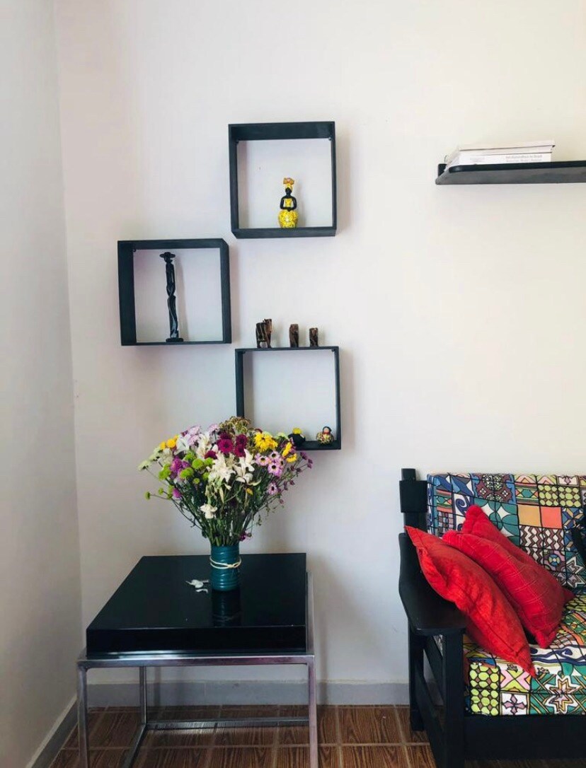 A corner of the living area features a colorful upholstered chair adorned with red and patterned accent pillows. A small glass table holds a vibrant bouquet of flowers. Above the table, minimalist black shelves display decorative items, enhancing the decor.