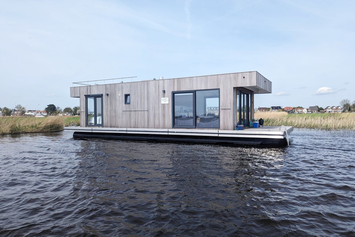 The houseboat displays a modern angular design with large windows, allowing for abundant natural light. It floats gently on calm waters, surrounded by lush greenery and reeds. The exterior is clad in wooden panels, contributing to its eco-friendly aesthetic.