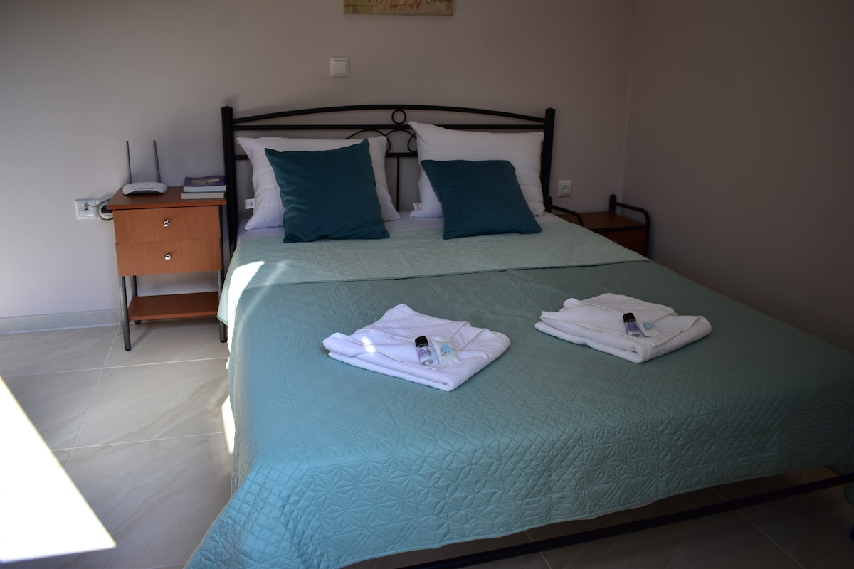 A well-appointed bedroom features a spacious bed with a light blue coverlet. Two accent pillows add a touch of color. Fresh towels are neatly arranged at the foot of the bed. A bedside table with books and a lamp is located beside the bed.
