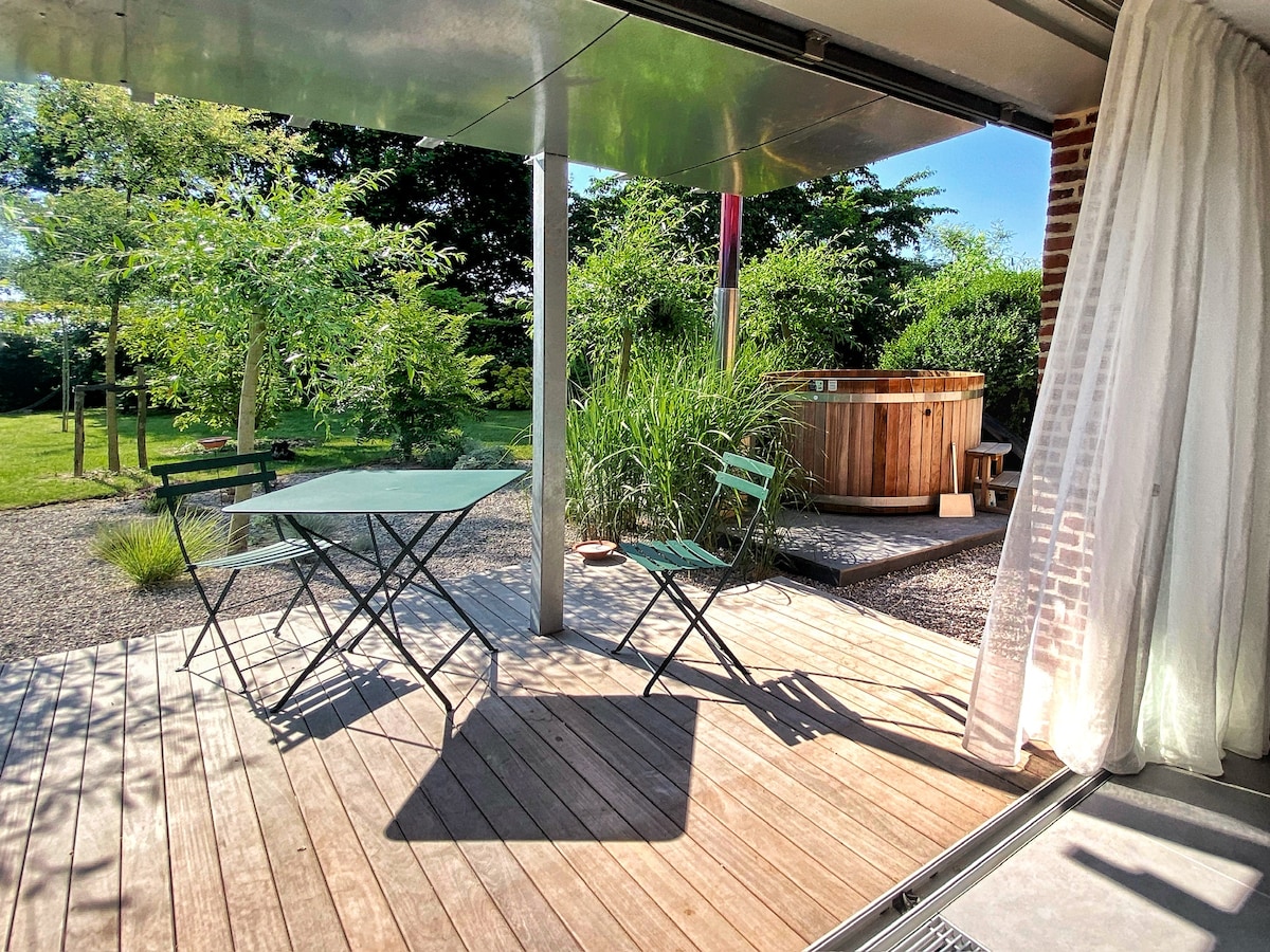A wooden terrace features a folding table and two green chairs, complemented by lush greenery in the background. A wooden hot tub is positioned nearby, surrounded by a tranquil garden space. Natural light filters through sheer curtains, enhancing the serene setting.