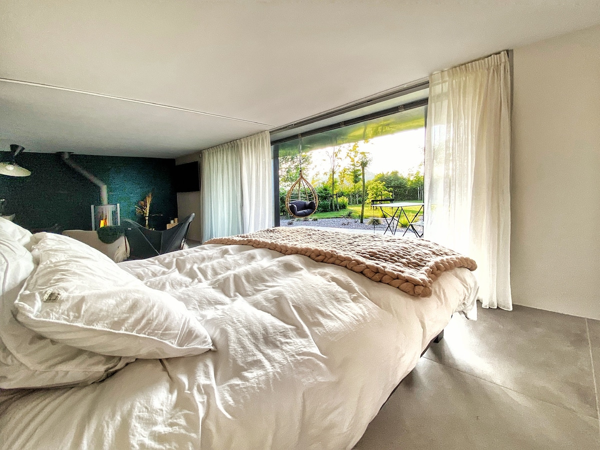 A spacious bed with layered white bedding is positioned in the foreground. Large glass windows allow natural light to fill the room, revealing a view of a garden. A hanging chair and a small outdoor table with chairs are visible on the terrace.