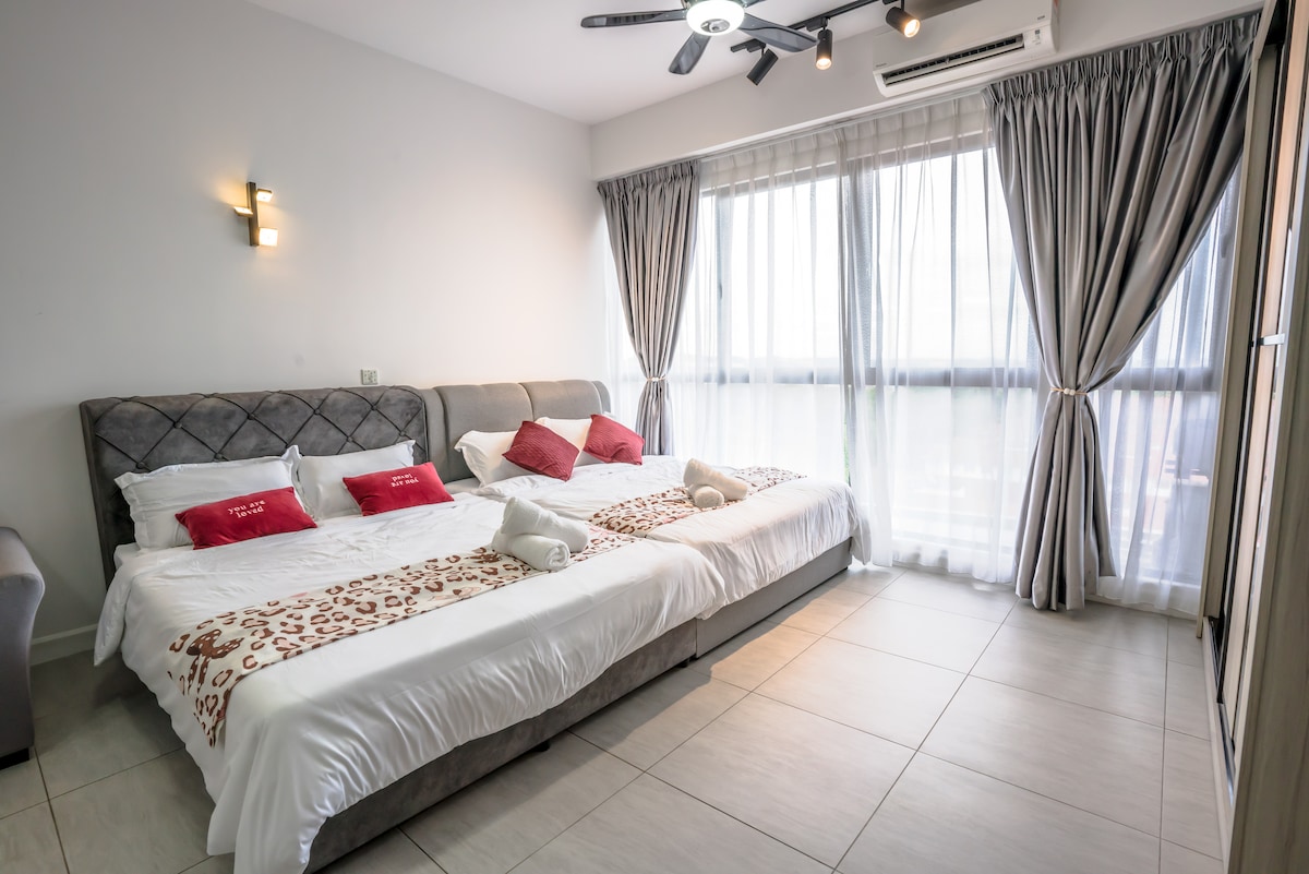 A well-lit bedroom features two spacious queen beds, accented with decorative pillows. Large windows are dressed in sheer curtains, allowing natural light to fill the room. A modern fan hangs from the ceiling, providing a comfortable atmosphere.