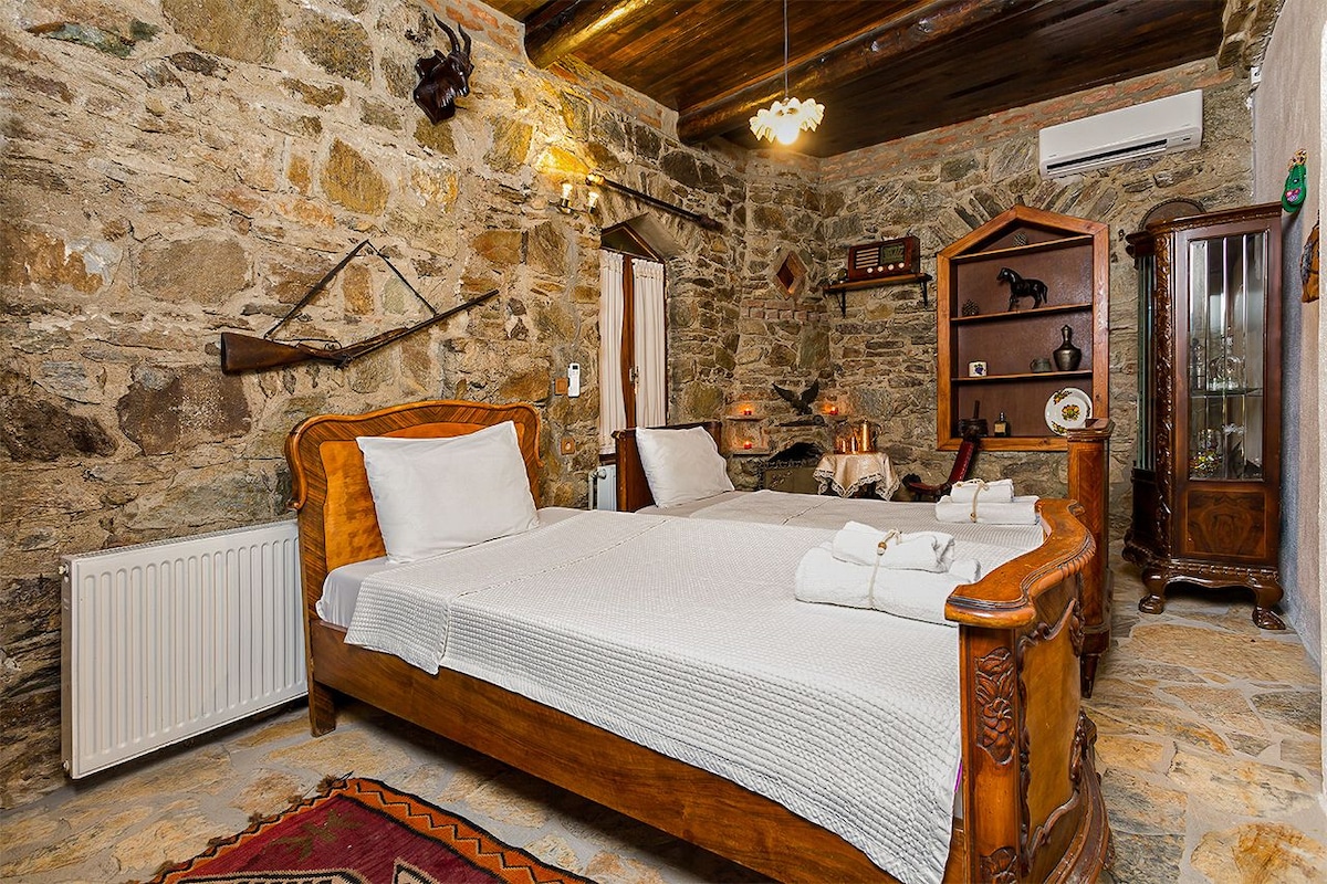 A rustic bedroom features stone walls and wooden beams, creating a traditional ambiance. Two spacious beds are elegantly framed in carved wood, accompanied by white linens. A glass cabinet and decorative shelves contain assorted decor items, adding character to the room.