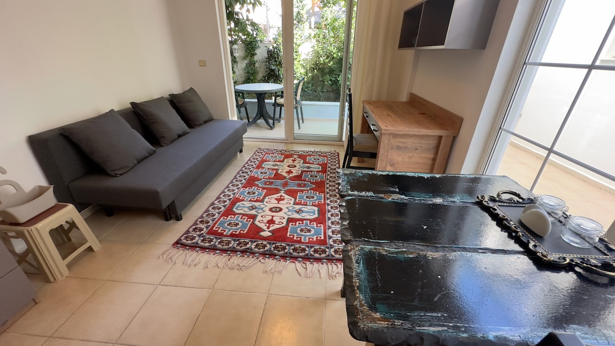 A simple living area features a dark sofa adorned with cushions, accompanied by a wooden desk and decorative items. A colorful patterned rug adds warmth to the tiled floor, while large glass doors lead to a garden view, enhancing the room's natural light.