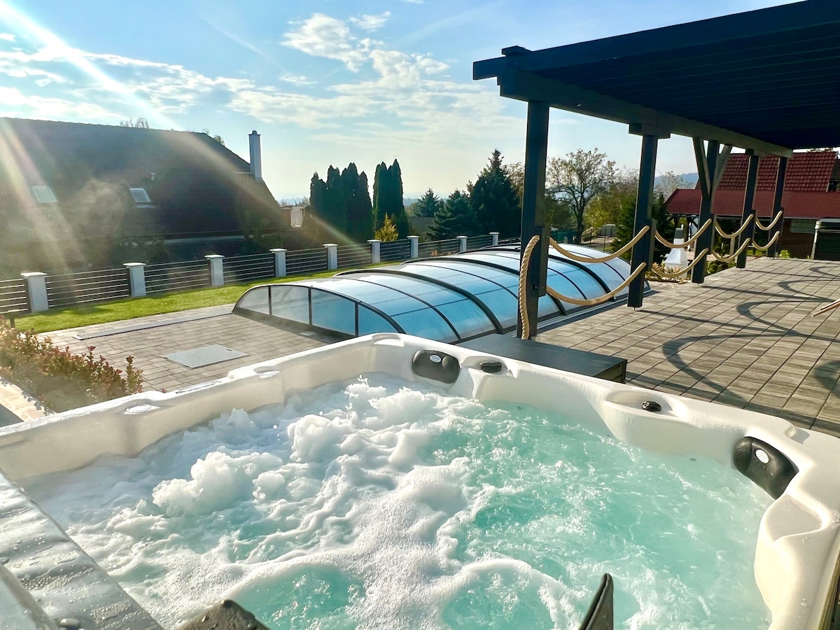 A hot tub with bubbling water is situated under a shaded area, offering views of the surrounding landscape. The patio features a sleek tiled surface, and a retractable cover is visible over an adjacent swimming pool, enhancing the outdoor space.