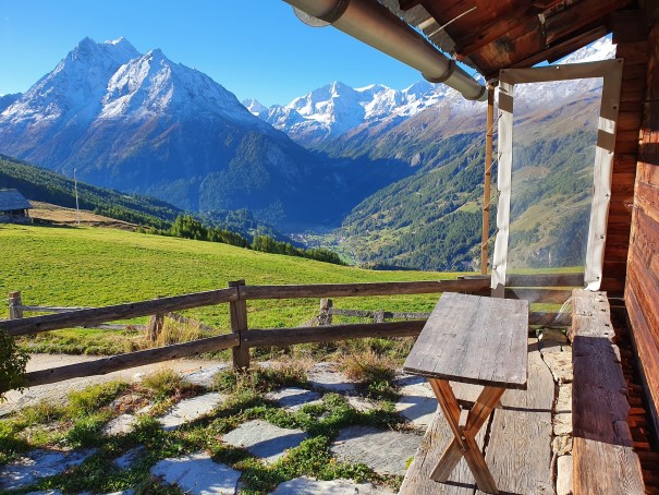 A wooden terrace provides a clear view of the surrounding mountains and valley. A small table is positioned on the terrace, inviting moments of relaxation. The rich green grass contrasts with the rugged mountain peaks in the background, under a bright blue sky.