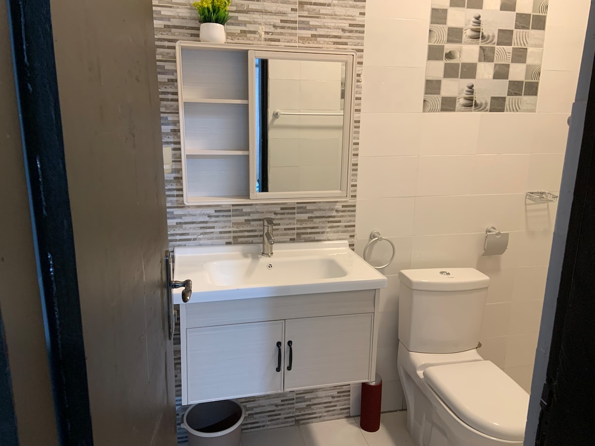 The bathroom features a modern vanity with a spacious sink and a large mirror above it. Textured wall tiles create a subtle contrast. A toilet is positioned opposite the vanity, with a roll of toilet paper and a small trash bin nearby.